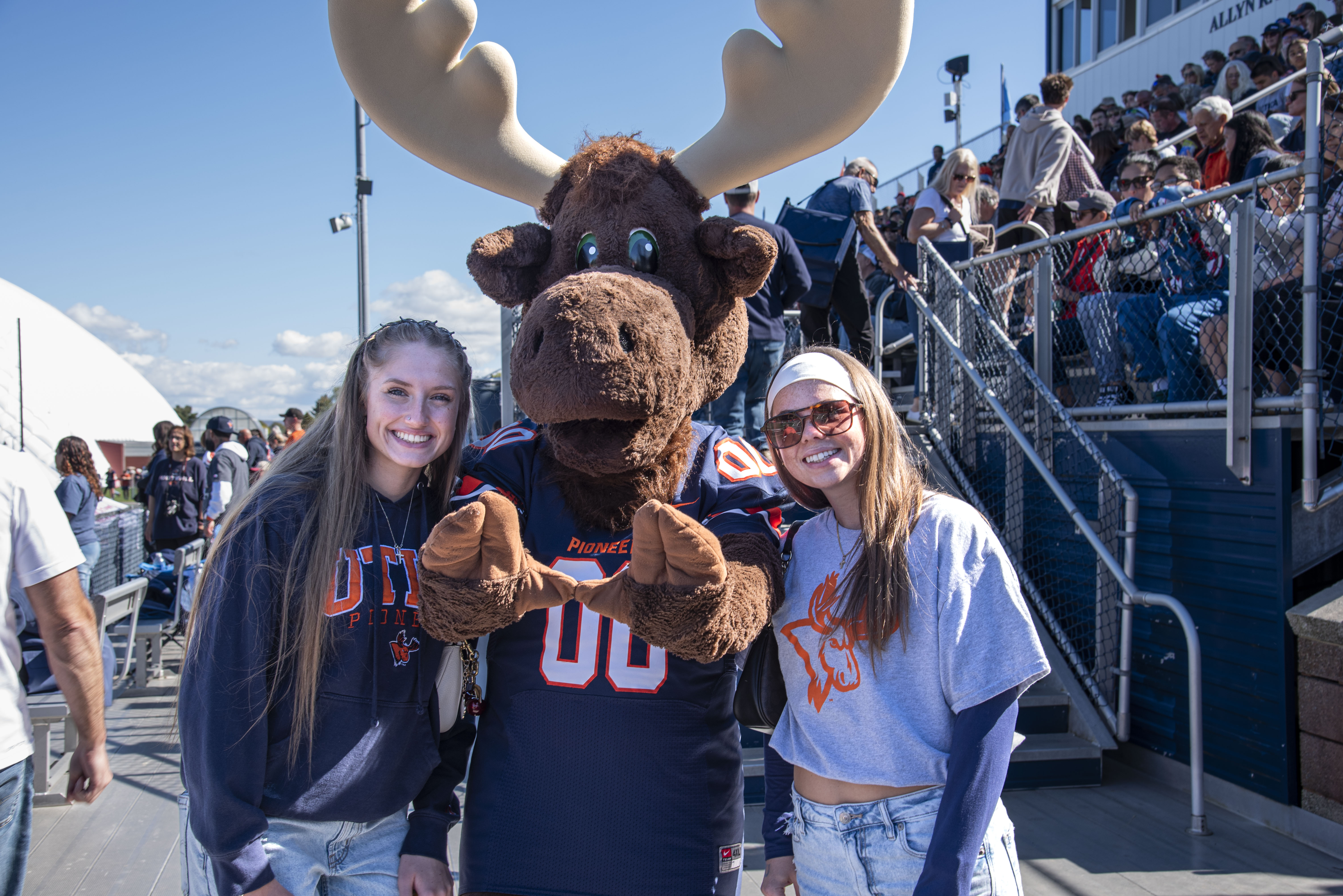 Two homecoming attendees stand on either side of Trax the Moose at the football game.
