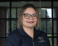 Mercedes Steele, in glasses and a blue Utica University polo shirt, stands in front of a window and smiles.