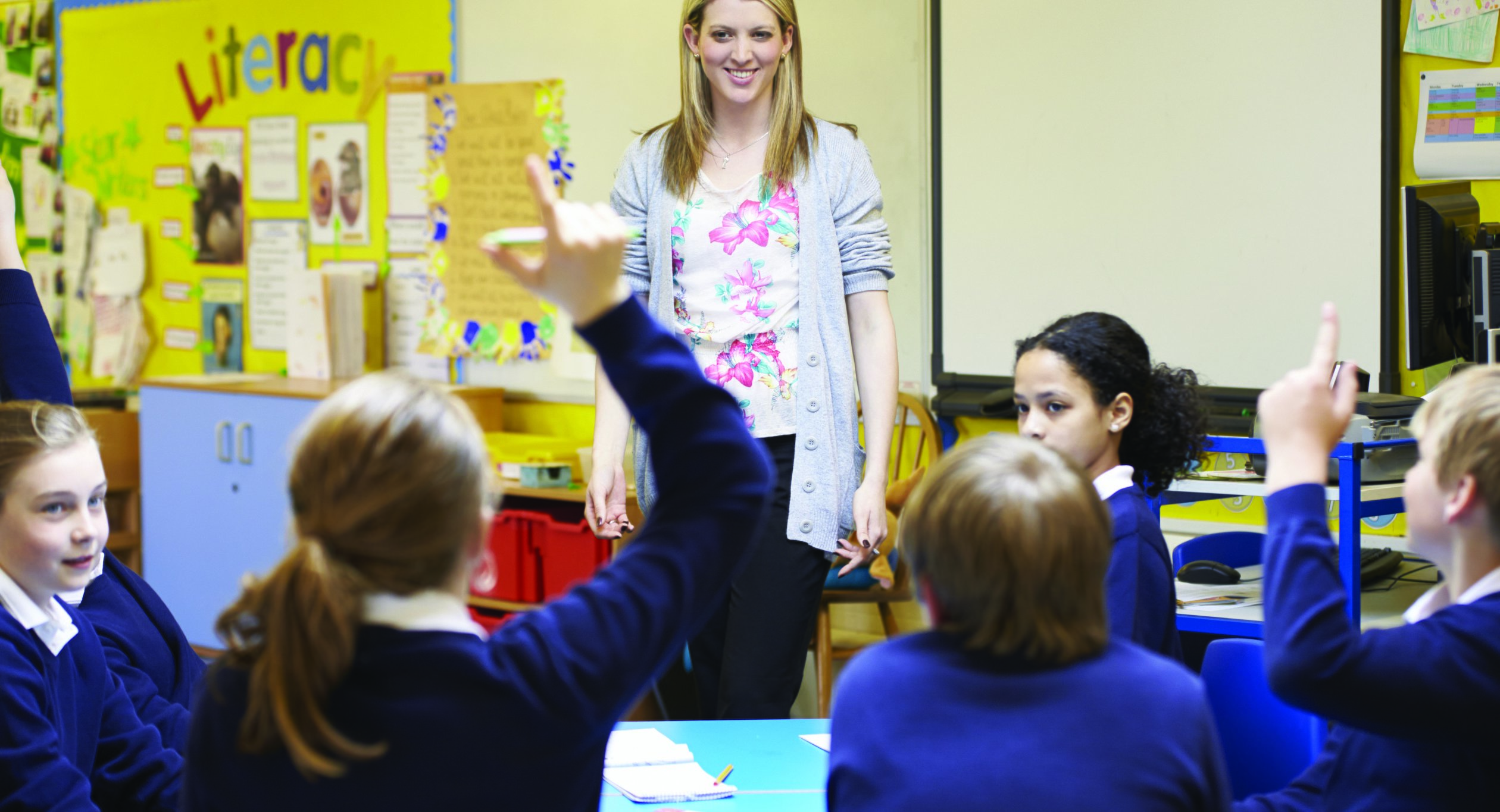 Teacher in a classroom with students raising their hands.