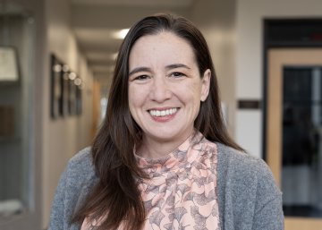 Deborah Pollack, with long brown hair, pink top and gray sweater, smiles at camera.