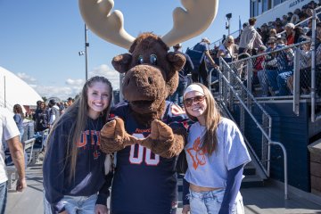 Two homecoming attendees stand on either side of Trax the Moose at the football game.