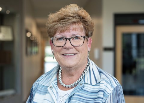 Tracy Pratt, with short hair and glasses, wearing a necklace and white and blue striped blazer, smiles at camera.