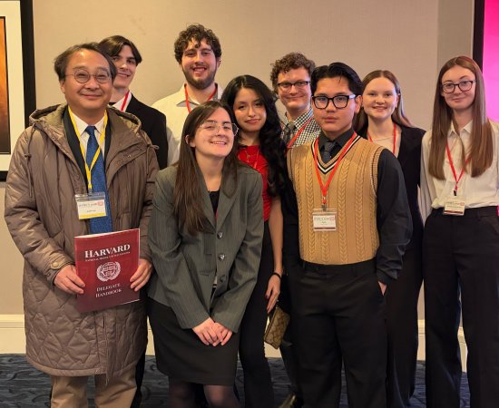 Students stand with professor, smiling, while holding a folder that says "Harvard" on it.