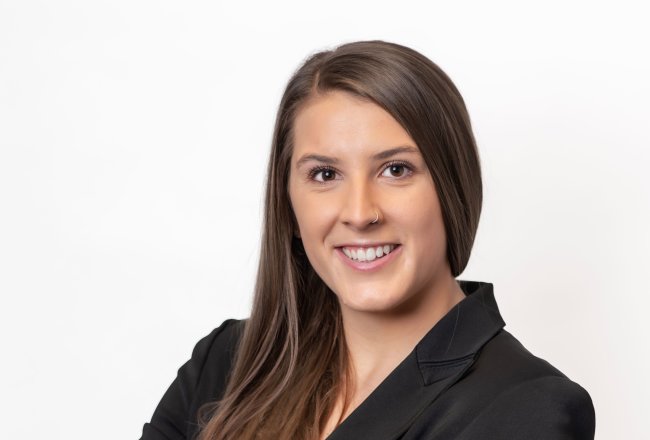 Madison Judge stands with arms folded in a black blazer, smiling at the camera against a white background.