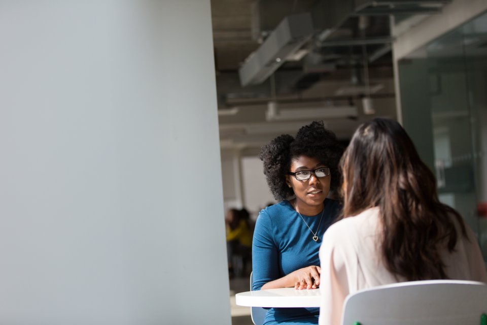 Woman at table speaking with another woman.