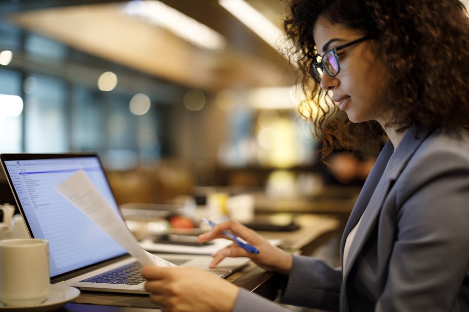 Woman in suit jacket and glasses holds a sheet of paper in one hand while typing on a laptop with the other hand.