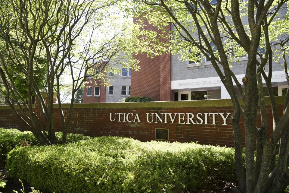 The words Utica University against a brick wall in front of a residence hall.